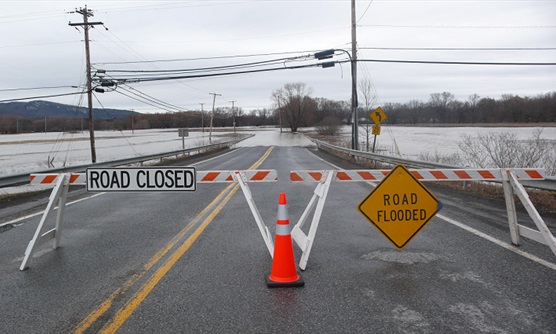 Flooing on a road and surrounding fields with road block barriers and "Flooded Road" sign - by L Heady